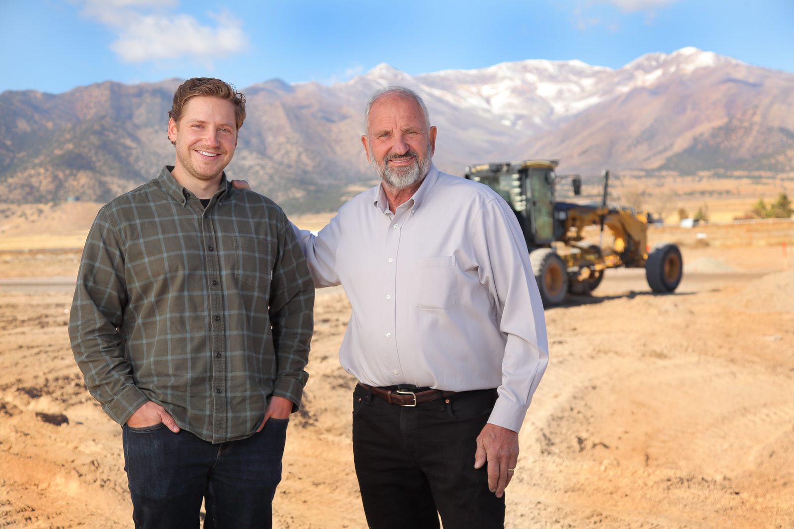 Garrett and Howard Schmidt standing together on a graded development site, with snow-capped Utah mountains and a road grader behind them